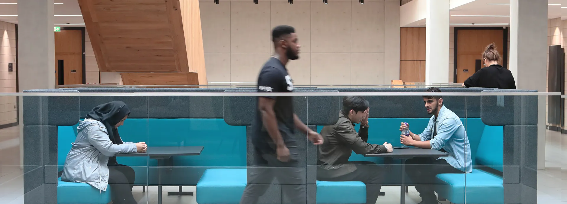 A modern indoor seating area with turquoise cushioned benches and glass railing, located in the University. Three people are seated at tables engaged in conversation or using devices, while one person walks past in the foreground, highlighting a dynamic and social environment.