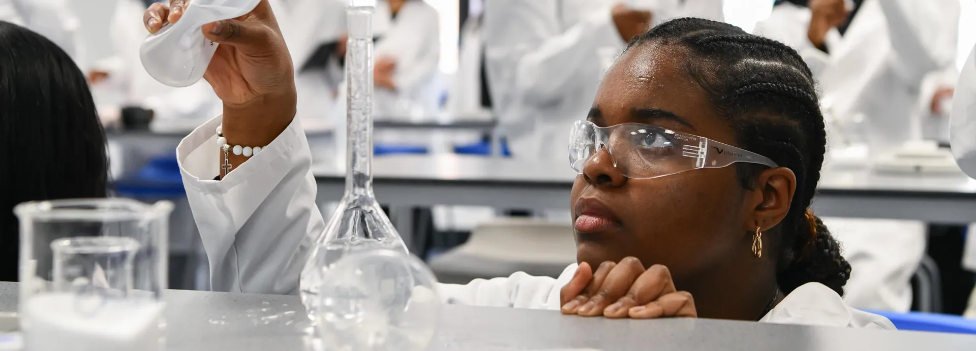 Photograph of students in a science laboratory conducting experiments with glassware and liquids. There is one student pouring liquid from a wash bottle into a graduated cylinder, surrounded by scientific equipment and peers in white lab coats.
