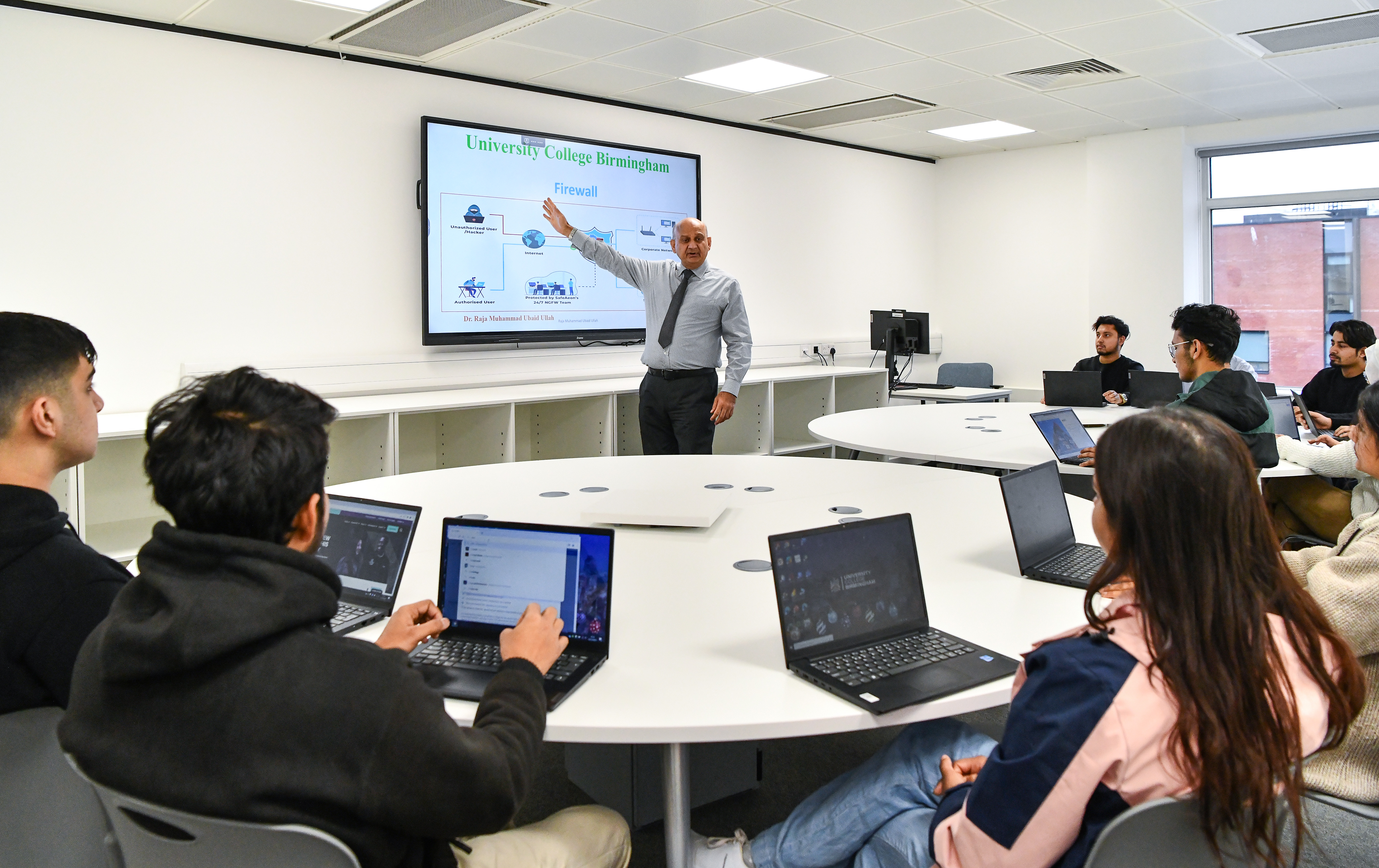 Male lecturer teaching a smaller group of students who are on laptops.