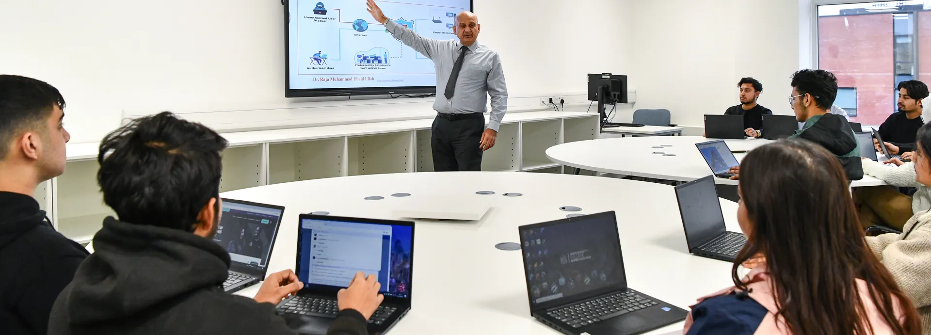 Male lecturer teaching a smaller group of students who are on laptops.