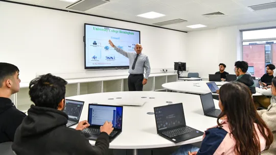 Male lecturer teaching a smaller group of students who are on laptops.