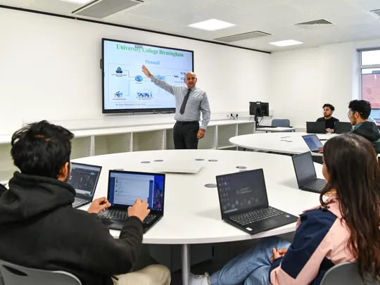 Male lecturer teaching a smaller group of students who are on laptops.