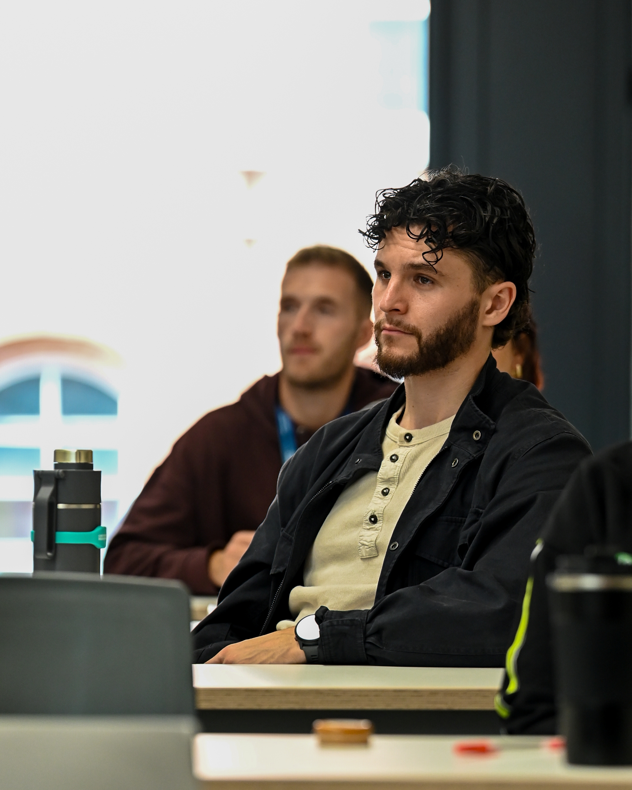 The man in the photo is a UCB alumus returning to start a social work apprenticeship, he is seated wearing a black jacket and cream top. He has short black curly hair and a beard.