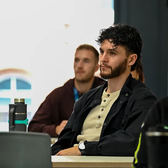 The man in the photo is a UCB alumus returning to start a social work apprenticeship, he is seated wearing a black jacket and cream top. He has short black curly hair and a beard.