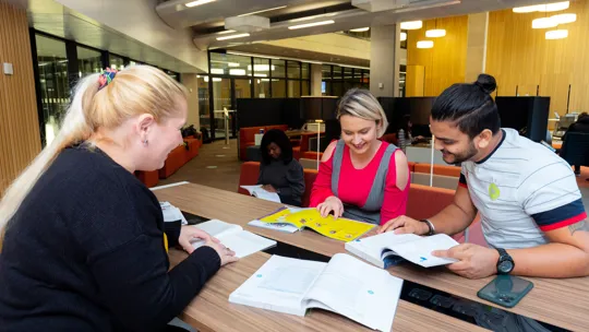 A group of four students studying together in the Postgraduate Suite in McIntyre House. Open textbooks, notebooks, and a smartphone are visible on the table, indicating a collaborative learning environment.