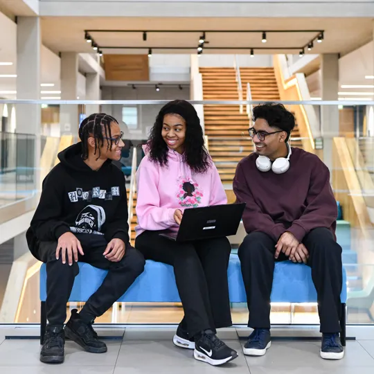 Three students sitting on a blue sofa in Moss House engaging with one another and a laptop on the middle students lap.