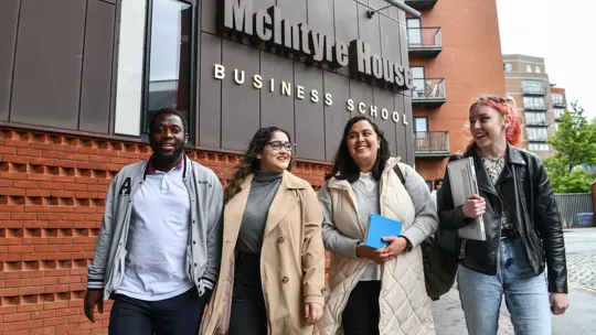 Group of students smiling walking in to campus with a Business school sign above them.