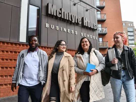 Group of students smiling walking in to campus with a Business school sign above them.