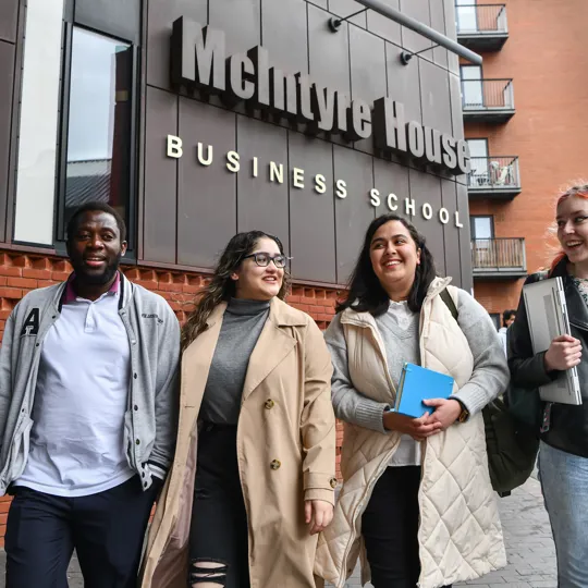 Group of students smiling walking in to campus with a Business school sign above them.