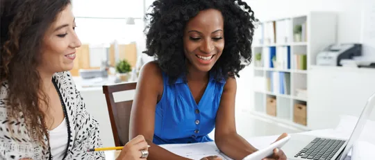 Two women in an office space working collaboratively on electronic devices.