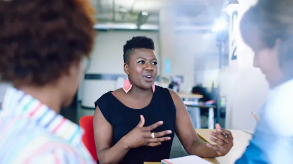 Three individuals engaging in a conversation with the middle person leading the meeting, wearing a black t-shirt and red earrings.