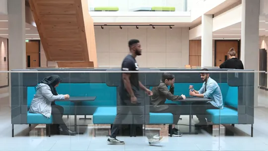 A modern indoor seating area with turquoise cushioned benches and glass railing, located in the University. Three people are seated at tables engaged in conversation or using devices, while one person walks past in the foreground, highlighting a dynamic and social environment.