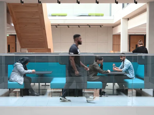 A modern indoor seating area with turquoise cushioned benches and glass railing, located in the University. Three people are seated at tables engaged in conversation or using devices, while one person walks past in the foreground, highlighting a dynamic and social environment.
