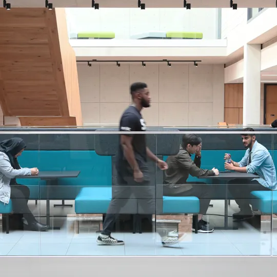 A modern indoor seating area with turquoise cushioned benches and glass railing, located in the University. Three people are seated at tables engaged in conversation or using devices, while one person walks past in the foreground, highlighting a dynamic and social environment.