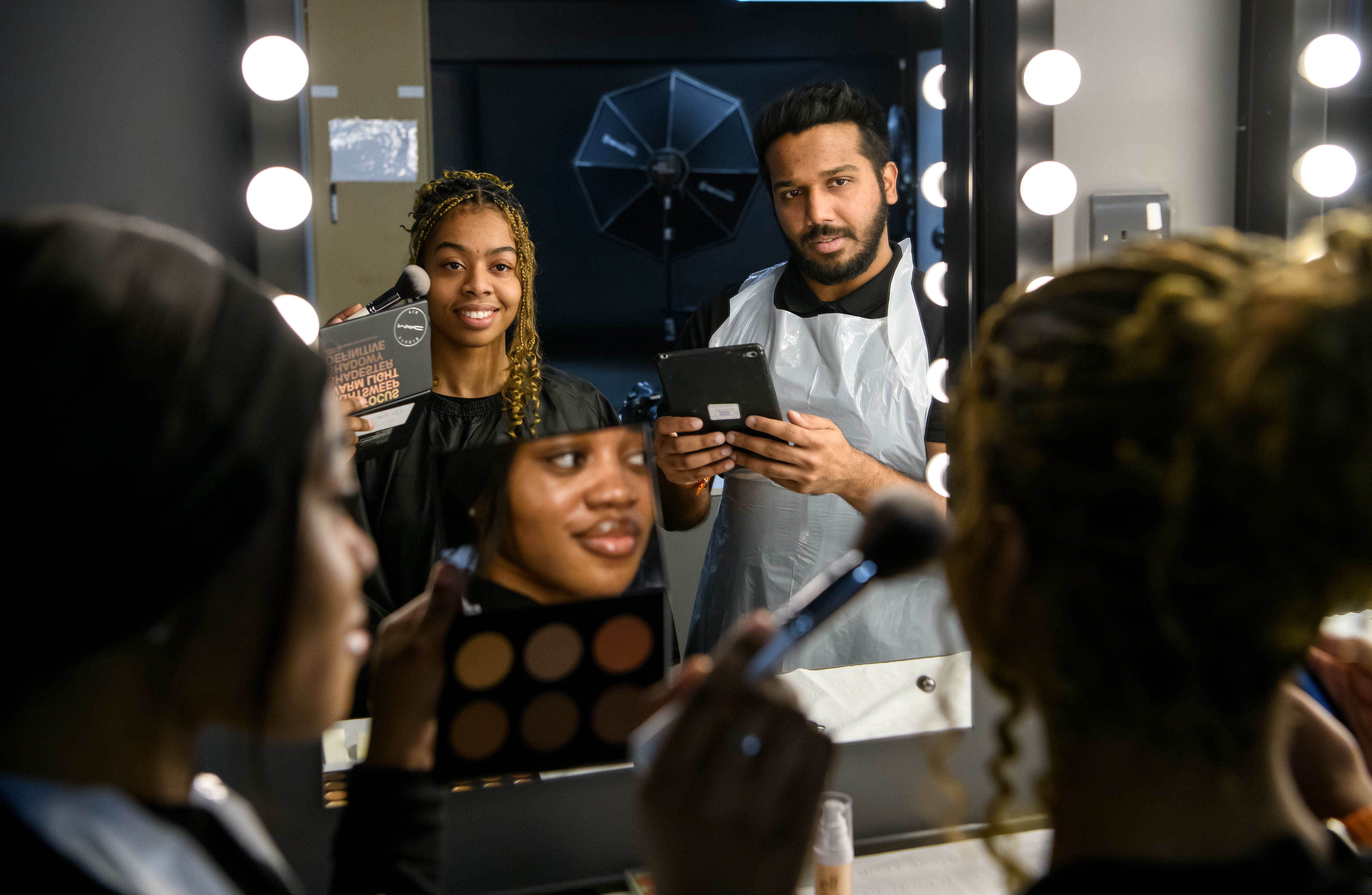 Female student aplying make-up to a female model.A male student is checking working using an ipad.