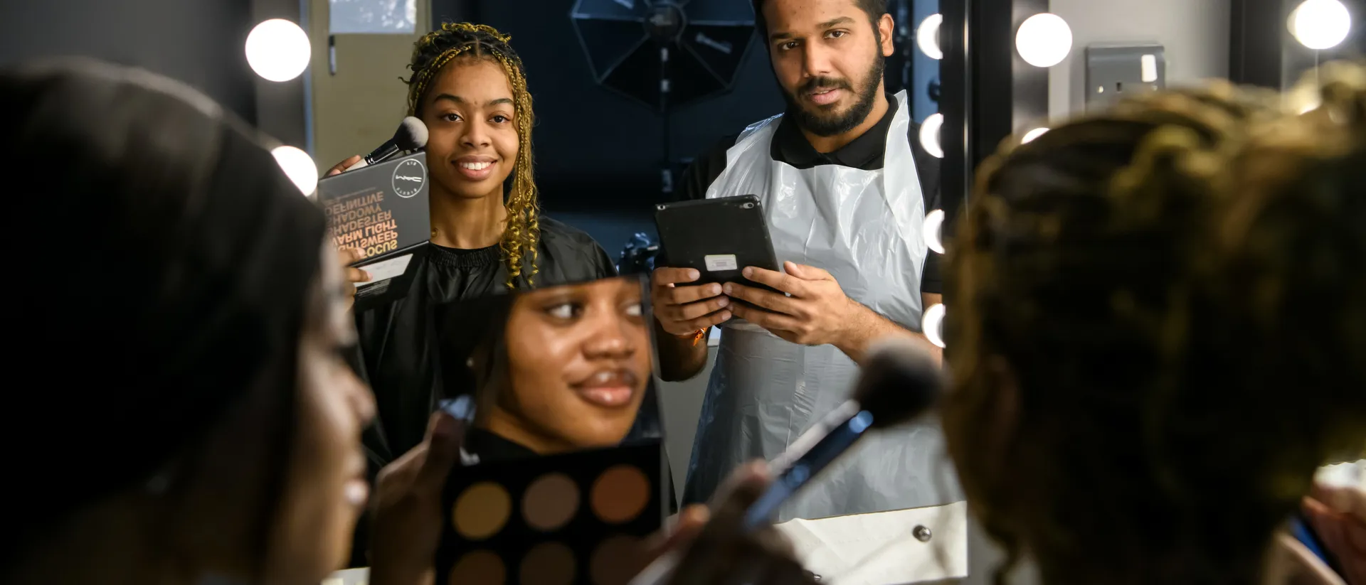 Female student aplying make-up to a female model.A male student is checking working using an ipad.