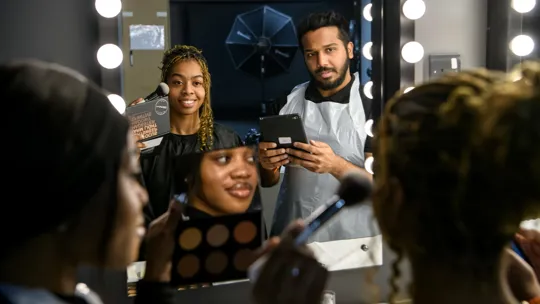 Female student aplying make-up to a female model.A male student is checking working using an ipad.