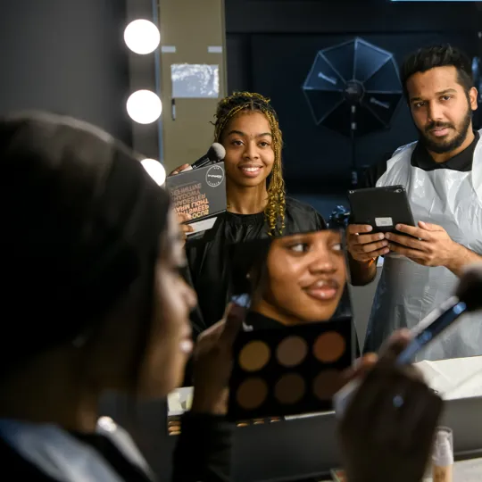 Female student aplying make-up to a female model.A male student is checking working using an ipad.