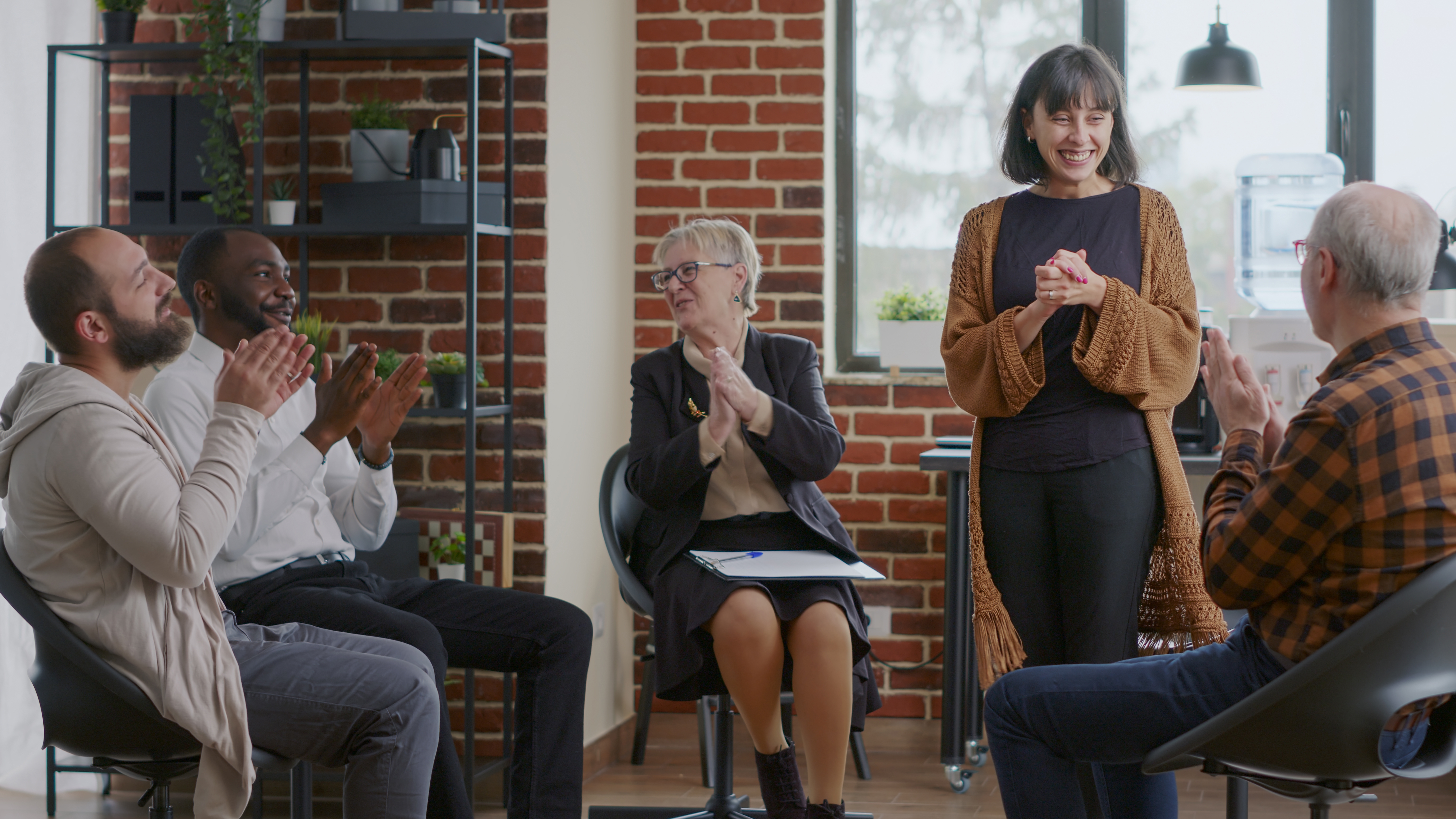 Group of adults in a support group setting with one person stood up and everyone clapping.