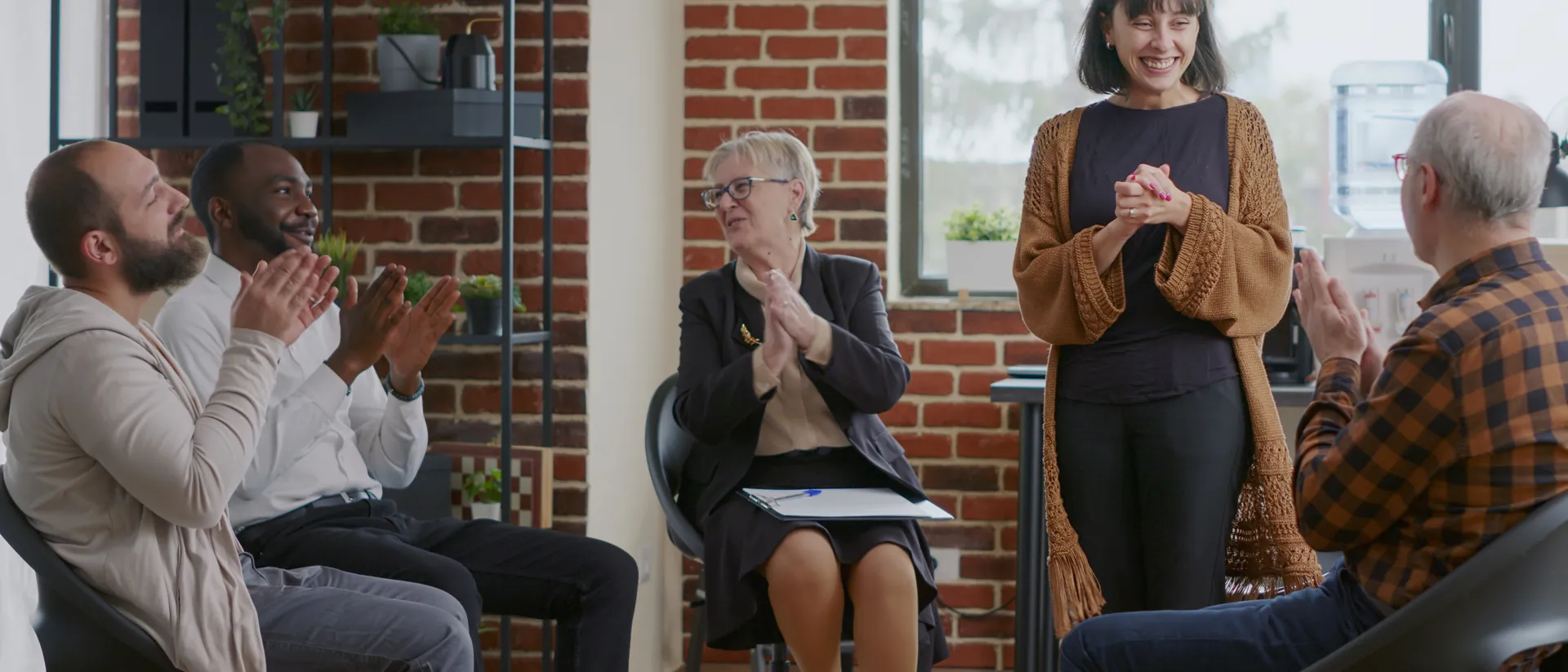 Group of adults in a support group setting with one person stood up and everyone clapping.