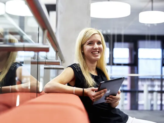 A woman smiling whilst holding an iPad in her hand seated on an orange Sofa.