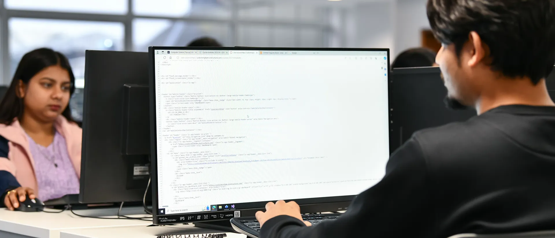 Male coding on a computer with a female in the background looking at her computer.