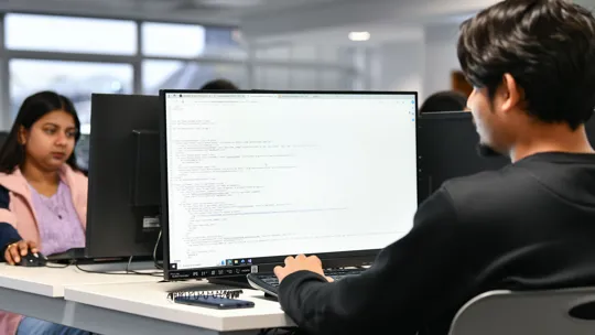 Male coding on a computer with a female in the background looking at her computer.