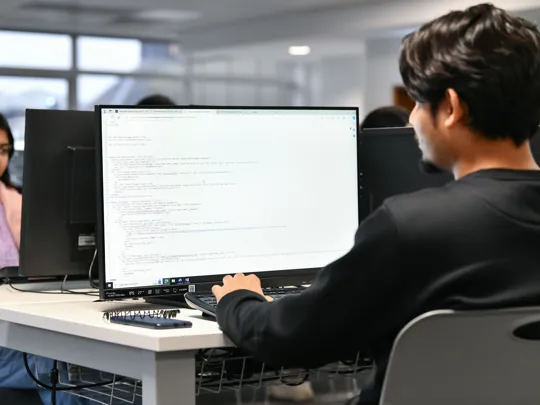Male coding on a computer with a female in the background looking at her computer.