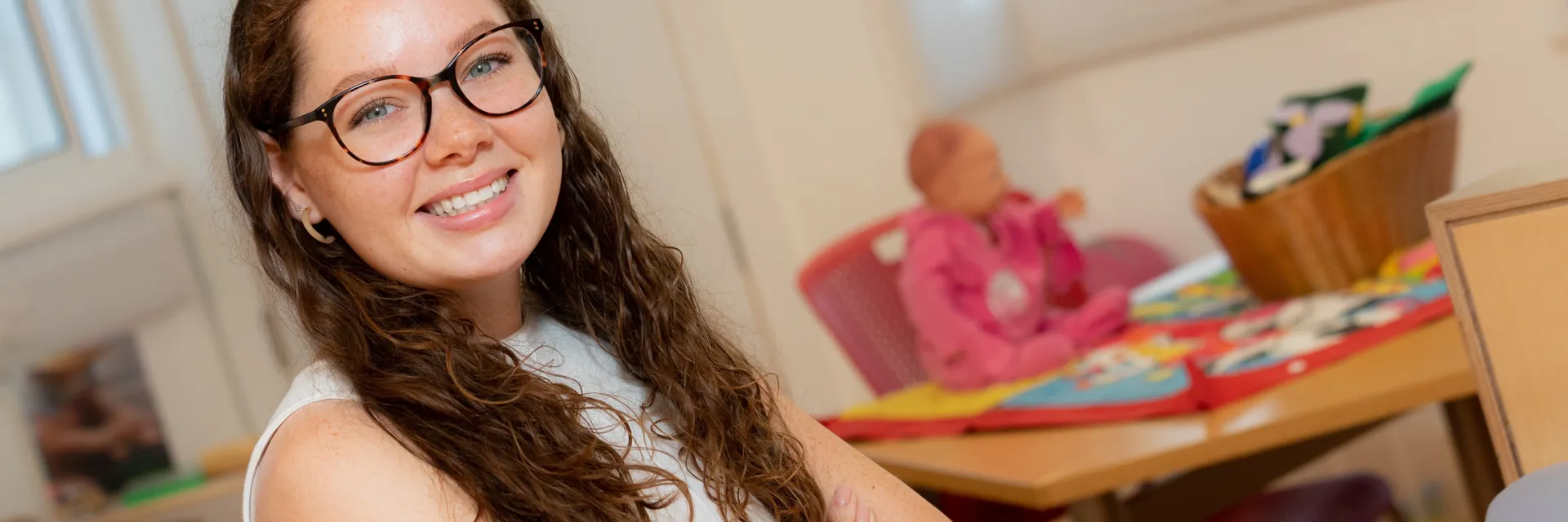 Childcare student smiling at the camera wearing black glasses inside a Nursery decorated classroom.