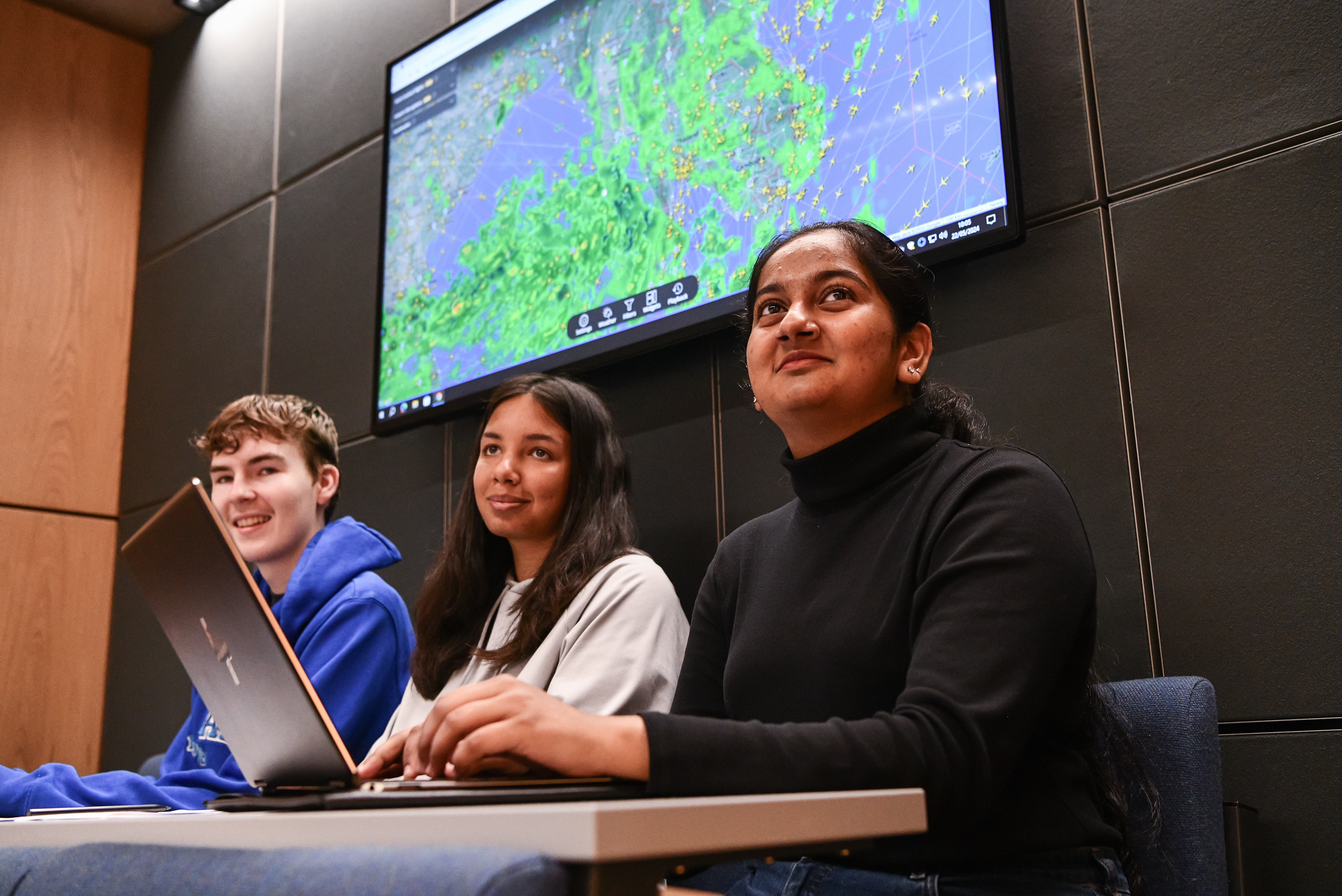 Three students sat with workbooks and one with a laptop smiling looking in the same direction. There is a large aviation map behind them.