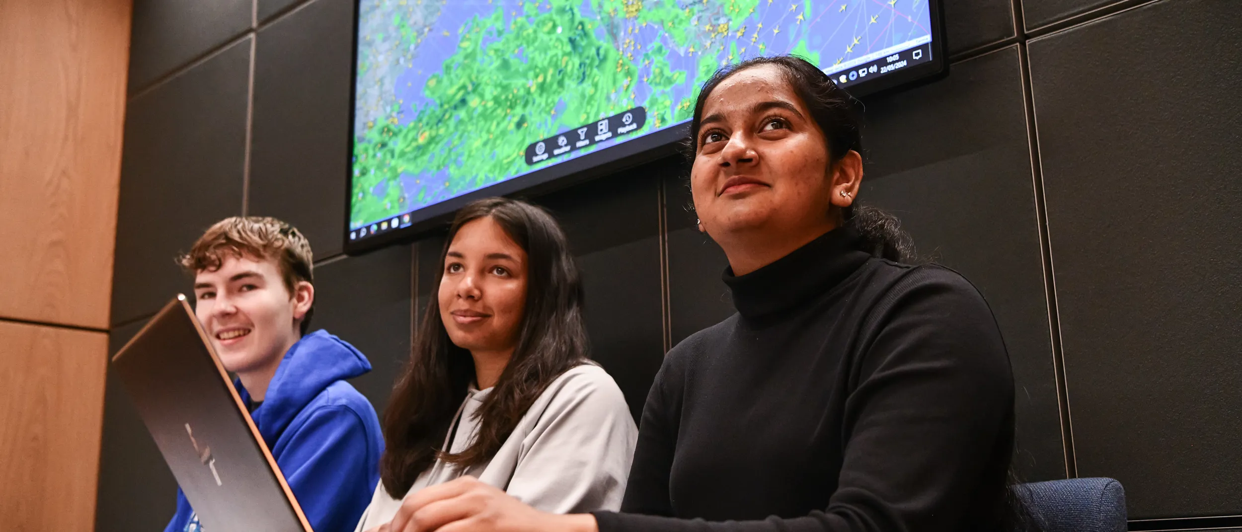 Three students sat with workbooks and one with a laptop smiling looking in the same direction. There is a large aviation map behind them.