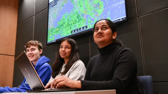 Three students sat with workbooks and one with a laptop smiling looking in the same direction. There is a large aviation map behind them.