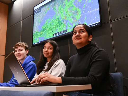 Three students sat with workbooks and one with a laptop smiling looking in the same direction. There is a large aviation map behind them.