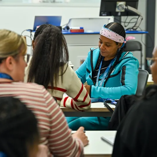 A student sits looking down at her work, she wears a bright blue tracksuit and a bandanna. She is one of the new students on UCB's social apprenticeship courses.