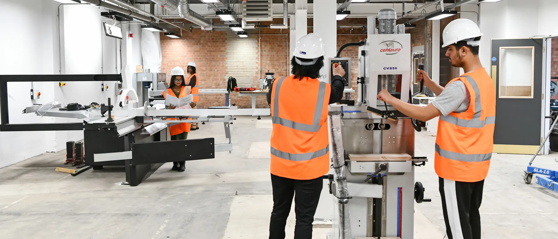 Construction students wearing helmets and orange high-visibility vests operating machinery inside the Construction Simulation space with exposed brick walls and concrete floors. The scene highlights teamwork and use of industrial grade equipment.