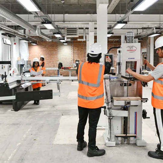Construction students wearing helmets and orange high-visibility vests operating machinery inside the Construction Simulation space with exposed brick walls and concrete floors. The scene highlights teamwork and use of industrial grade equipment.