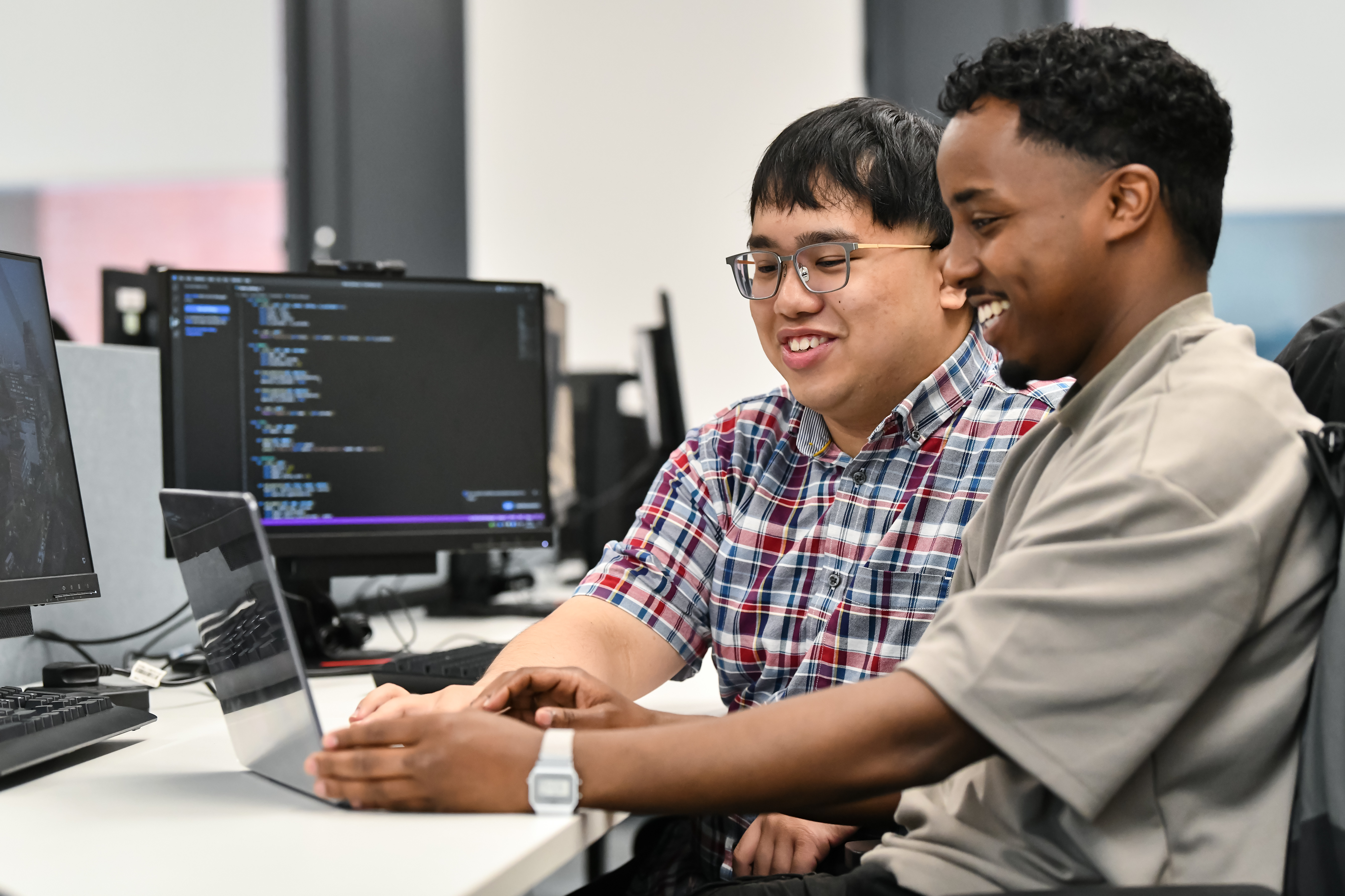 Two male students working on a desktop computer with a screen showing code. They are smiling.
