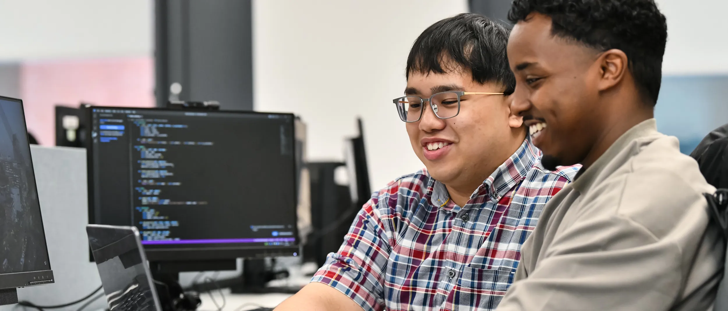 Two male students working on a desktop computer with a screen showing code. They are smiling.