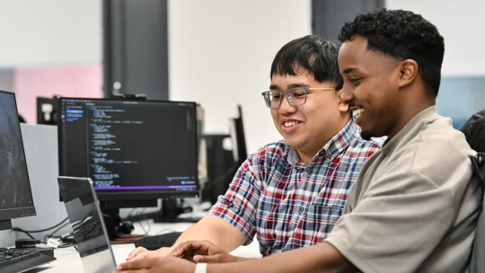 Two male students working on a desktop computer with a screen showing code. They are smiling.