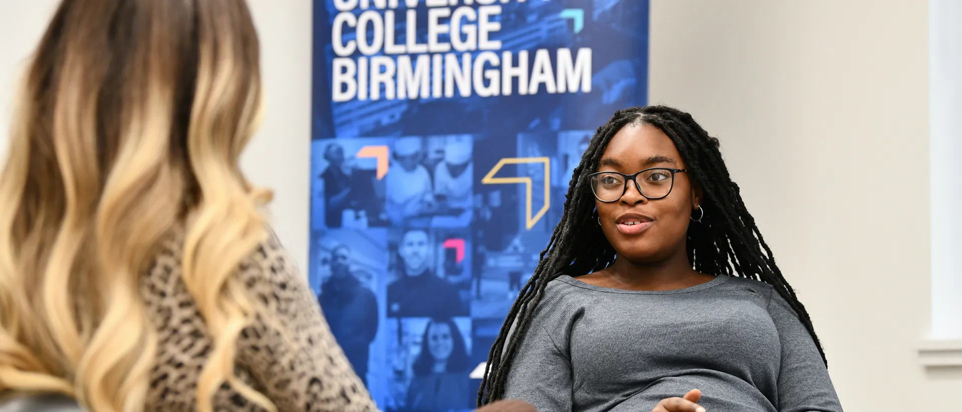 Two students sat talking with a banner that says we are University College Birmingham behind them.