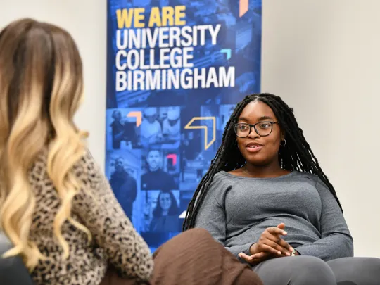 Two students sat talking with a banner that says we are University College Birmingham behind them.