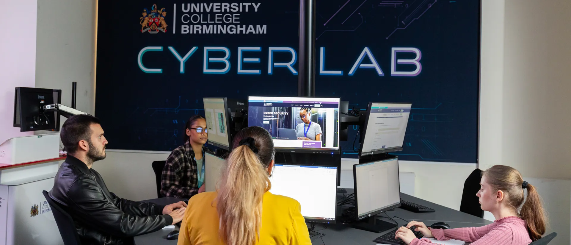 A group of students working on computers in the Cyber Lab at University College Birmingham. The room features multiple monitors, with a large illuminated wall sign reading 'CYBER LAB.' The students appear engaged, with one person taking notes from a book and others interacting with their screens.